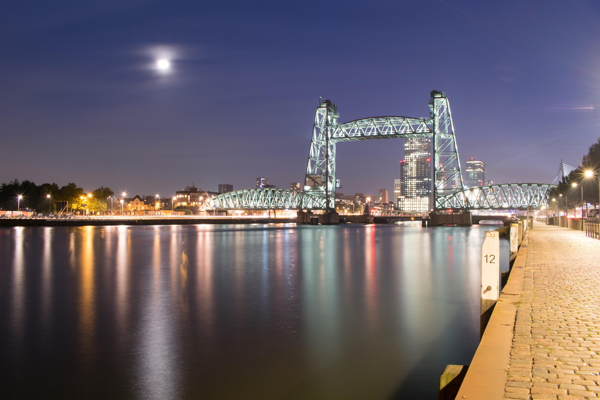 A steel bridge illuminated at night with a city skyline in the background and a full moon in the sky.